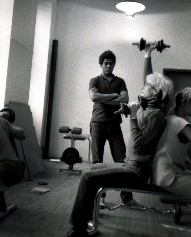 black and white photo of Andy Warhol lifting weights with Jean-Michel Basquiat looking on and a weight set in the background