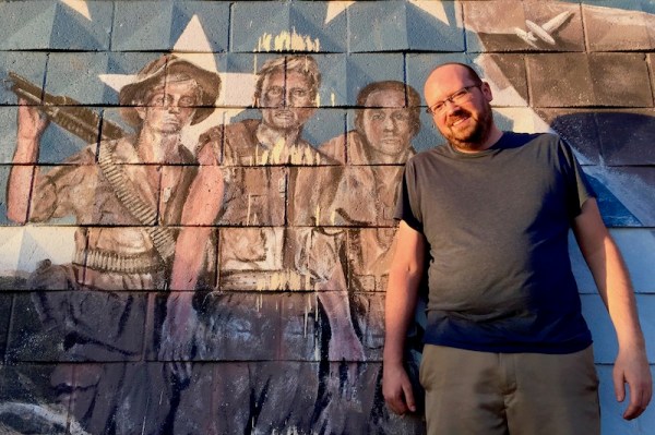 Poet Scott Silsbe standing in front of faded mural of Viet Nam veterans, Pittsburgh, PA