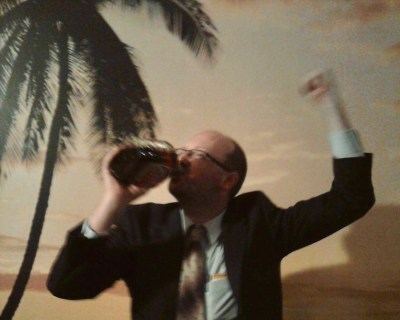 author Scott Silsbe dressed in suit and tie, drinking whiskey from a bottle in front of a wallpaper beach scene