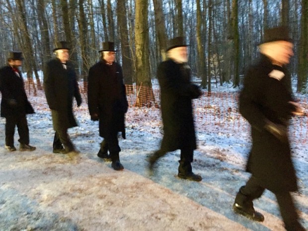Five men dressed in black pants, long coats, and top hats walk through winter woods