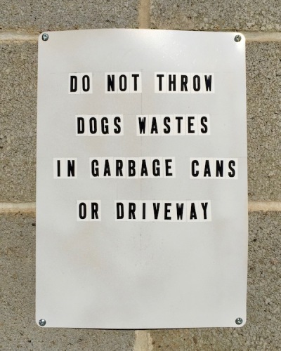 Sign posted on garage wall reading "Do not throw dogs wastes in garbage cans or driveway,"