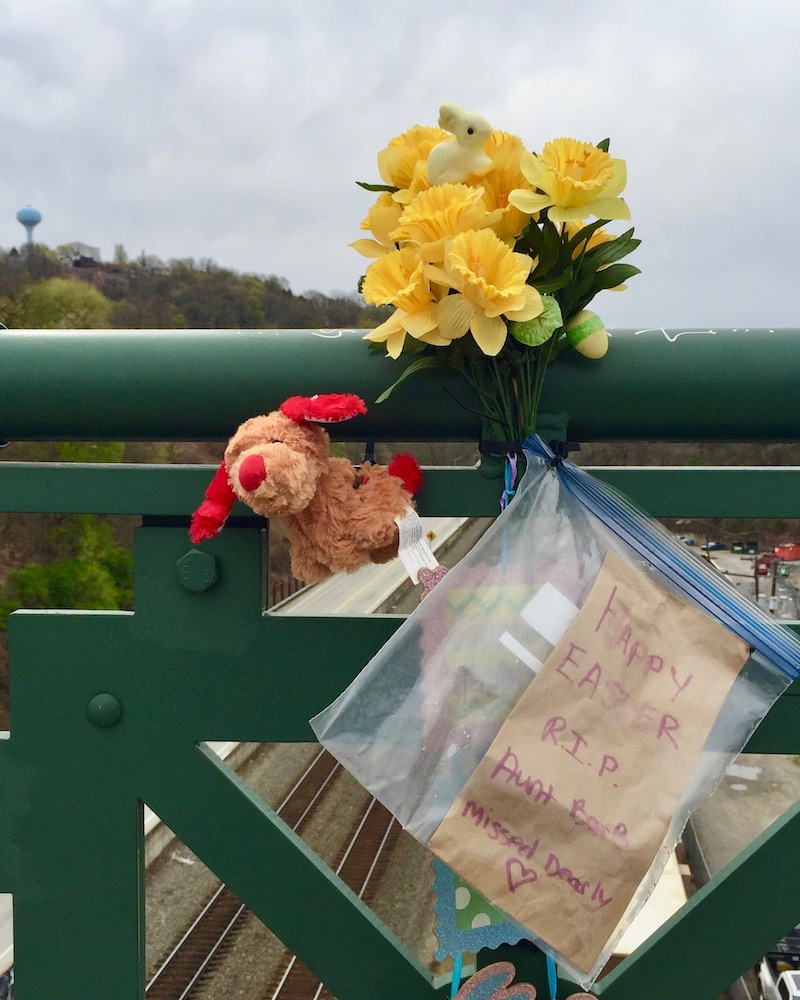memorial display with flowers, stuffed animal, and letter on bridge railing
