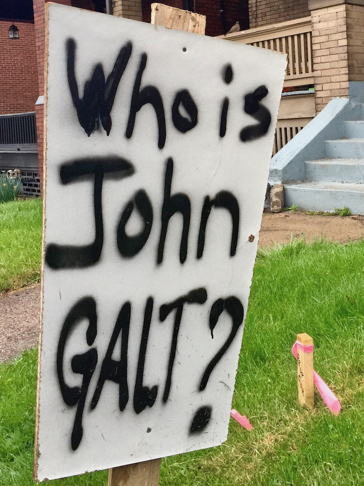 sign in front yard with spray-painted message "Who is John Galt?"