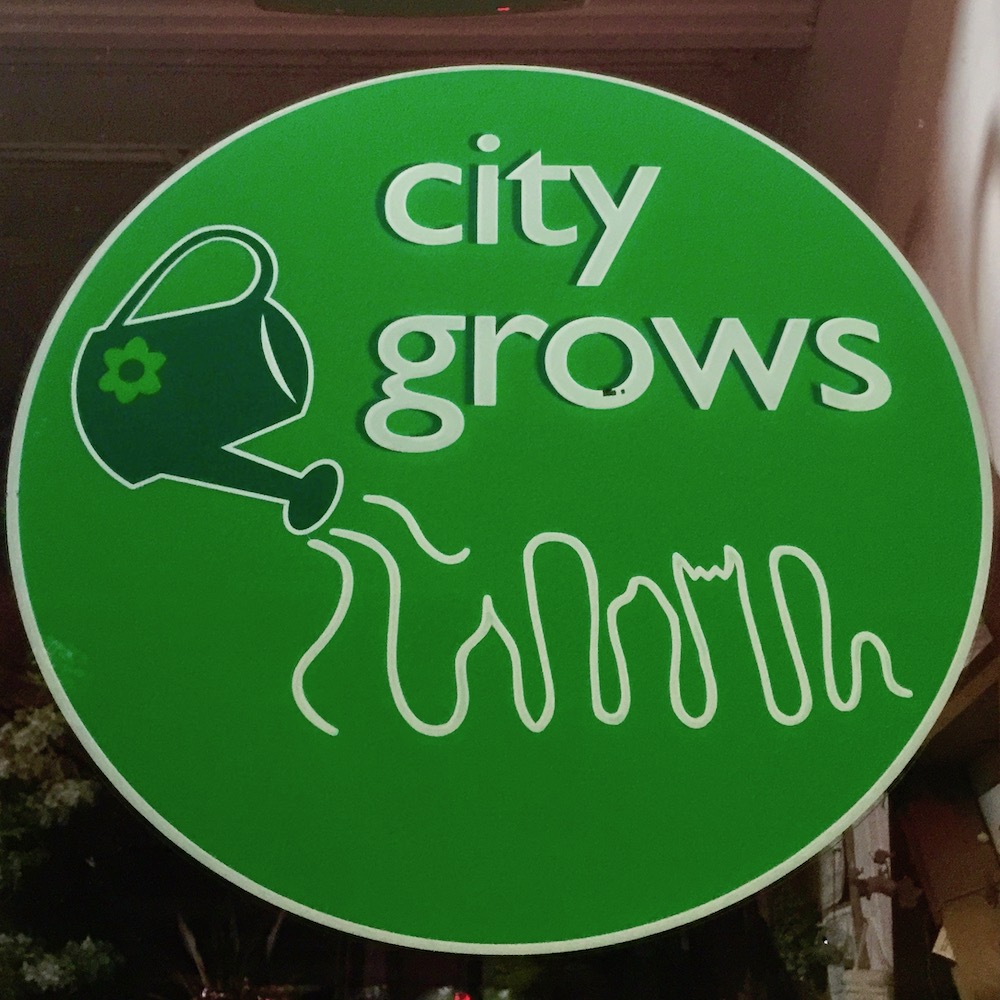 window sign for City Grows featuring downtown Pittsburgh buildings as growing grass