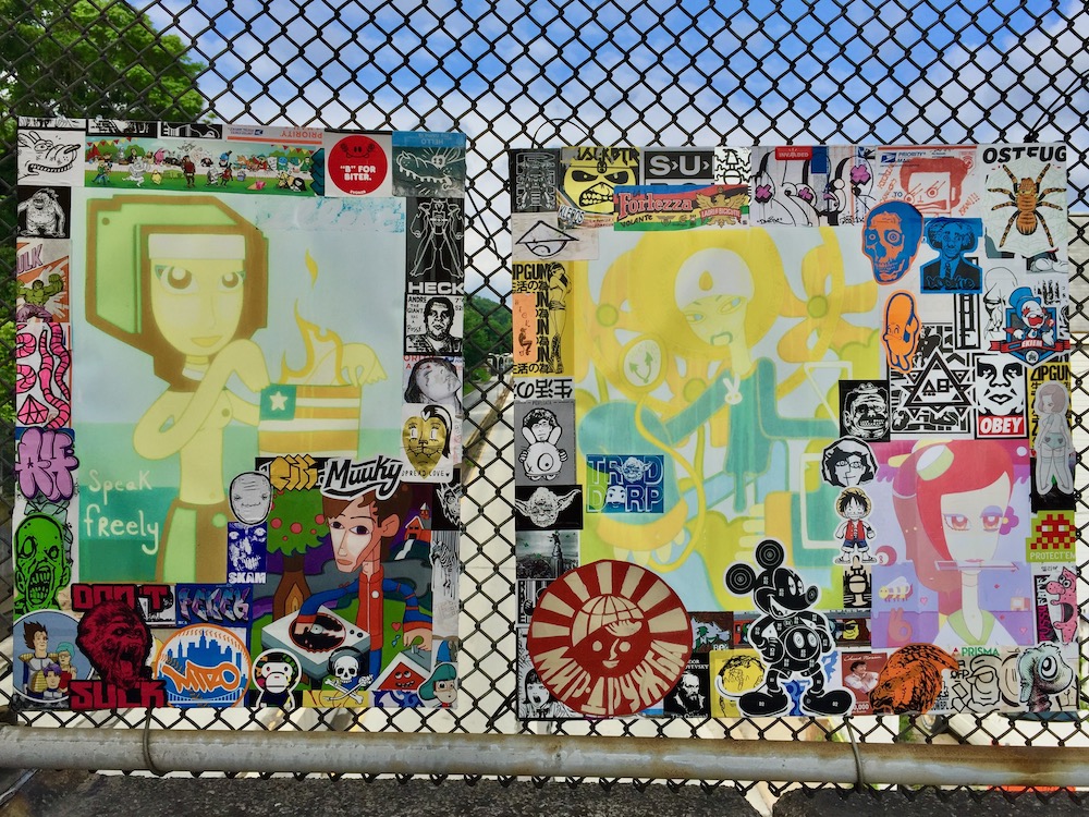 colorful collage of cartoon images, stickers, advertiments hung on chain link fence