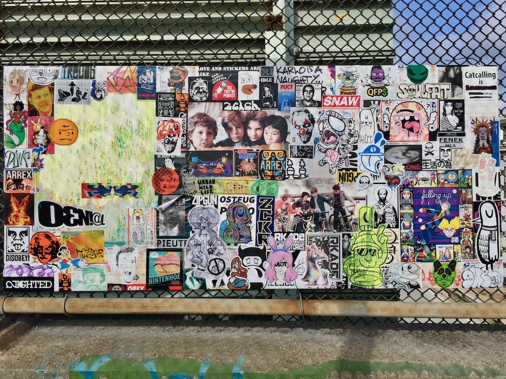 colorful collage of cartoon images, stickers, advertiments hung on chain link fence