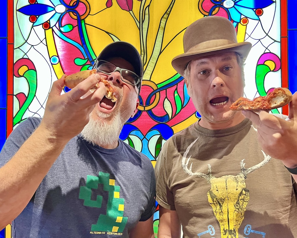 two men holding slices of pizza in front of a stained-glass window
