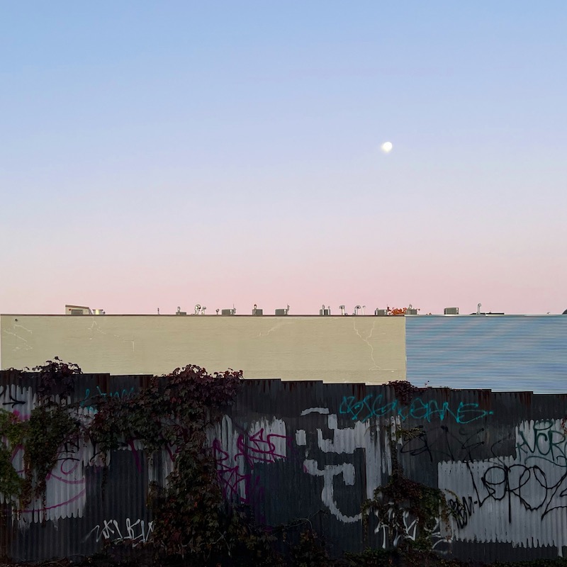 looking over graffiti-covered fence and large building to moon in sky
