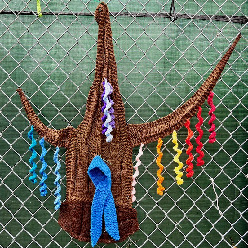 fiber art tree with colored streamers attached to chain link fence