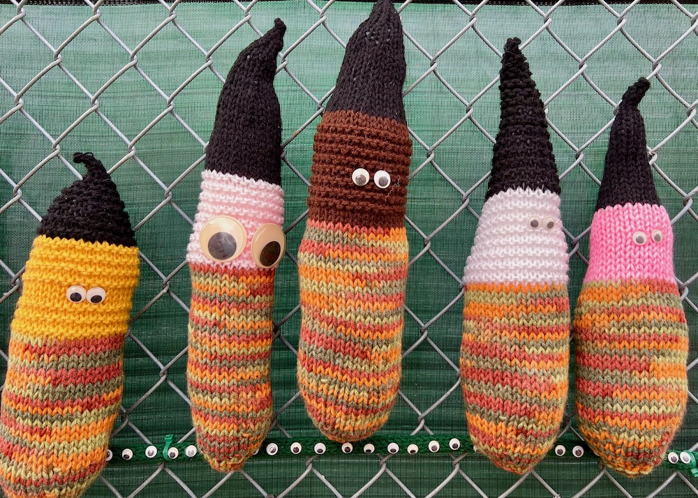 five crocheted figures with black hats and googly eyes attached to chain link fence
